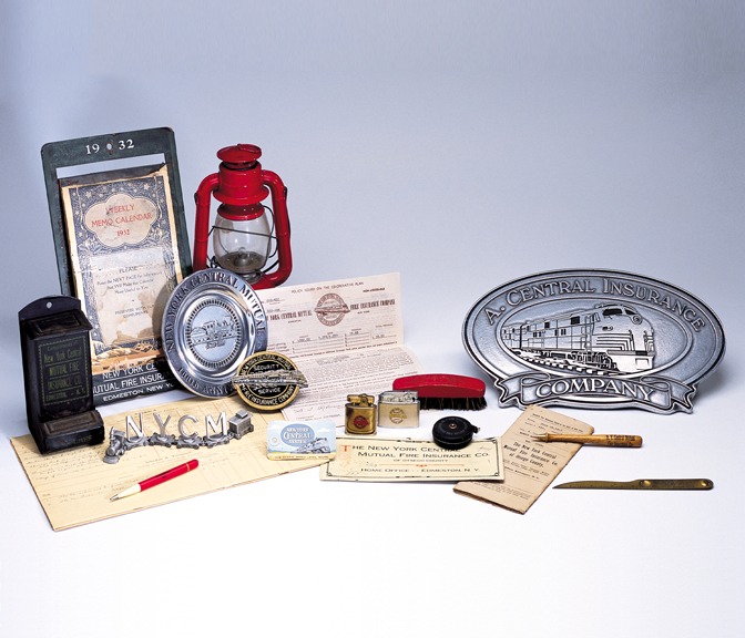 A small collection of NYCM Insurance's historical branded items: silver plates, knife, pen, red train light, train logo items, papers.